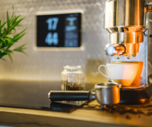 A modern espresso machine with a steaming cup of coffee beneath it. A digital clock is visible in the background, along with a small jar of coffee beans and a tamper tool.