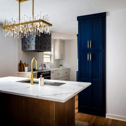 Modern kitchen interior with a white marble countertop island featuring a gold faucet, crystal chandelier, and a blue cabinet pantry with gold handles. The background includes a stylish black range hood with floral patterns and white cabinetry.