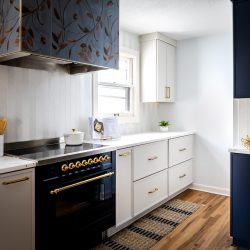 Modern kitchen with a black and gold stove, floral-patterned range hood, and white cabinetry. Decorative items, cutting boards, and a plant add style to the countertop.