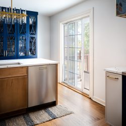 Modern kitchen with gold faucet, wooden and navy blue cabinetry, stainless steel dishwasher, and sliding glass door leading to an outdoor deck area.